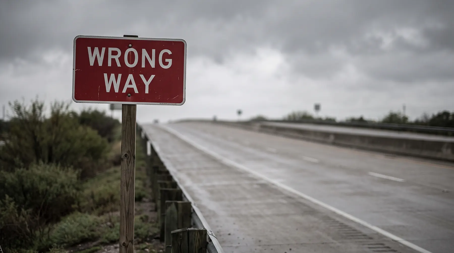 Red and white WRONG WAY road sign against an overcast sky above an empty highway off-ramp