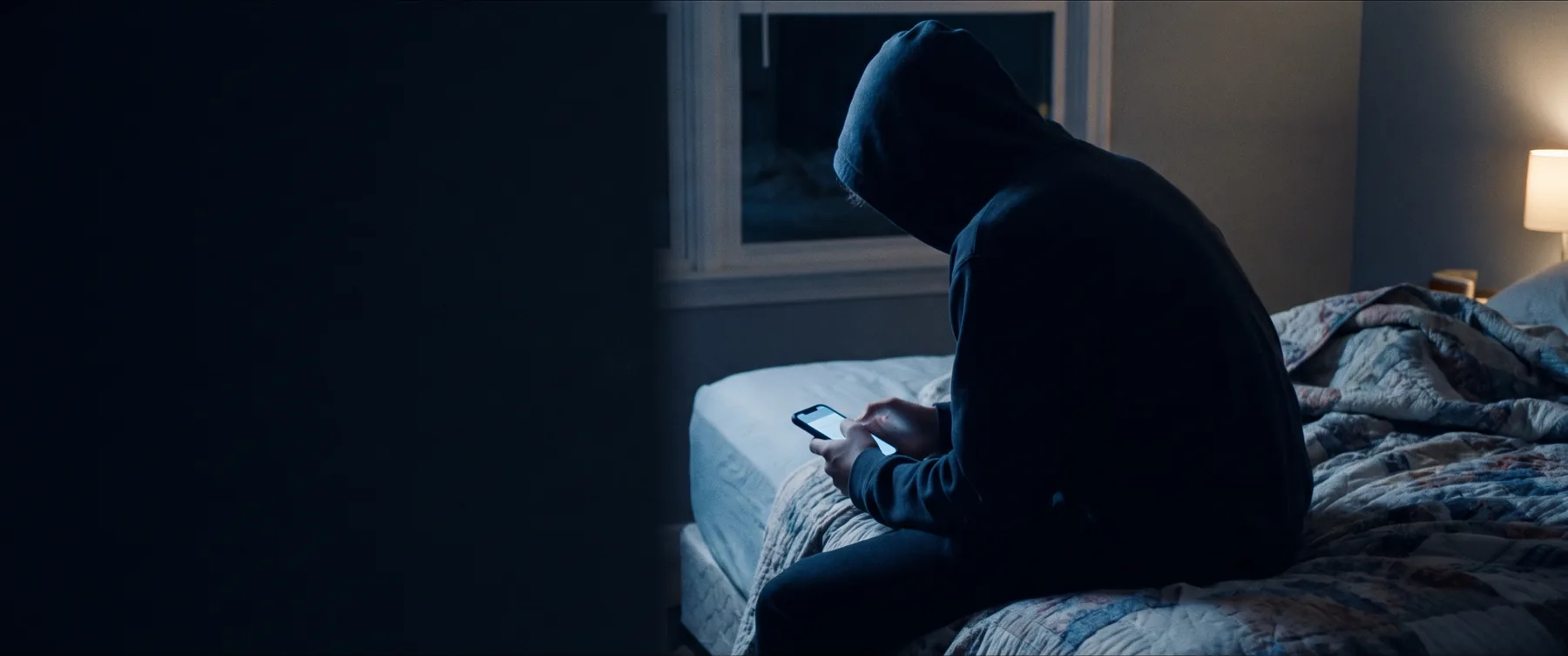 Teenager sitting on a bed in a dark room, lit only by a phone screen